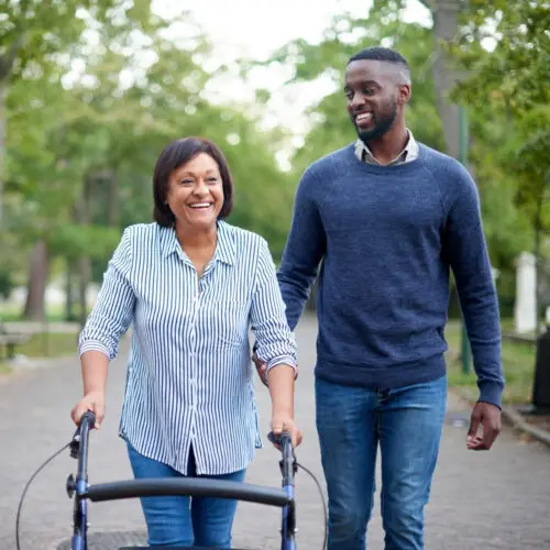 a young adult man smiles at an older woman using a walker as they walk down the street