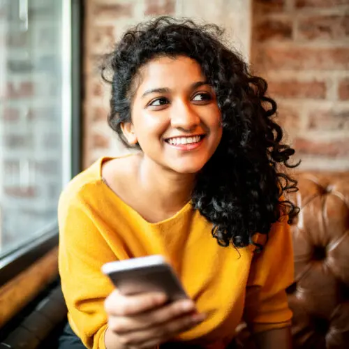 woman sitting by a window smiling at something off camera while holding a smartphone