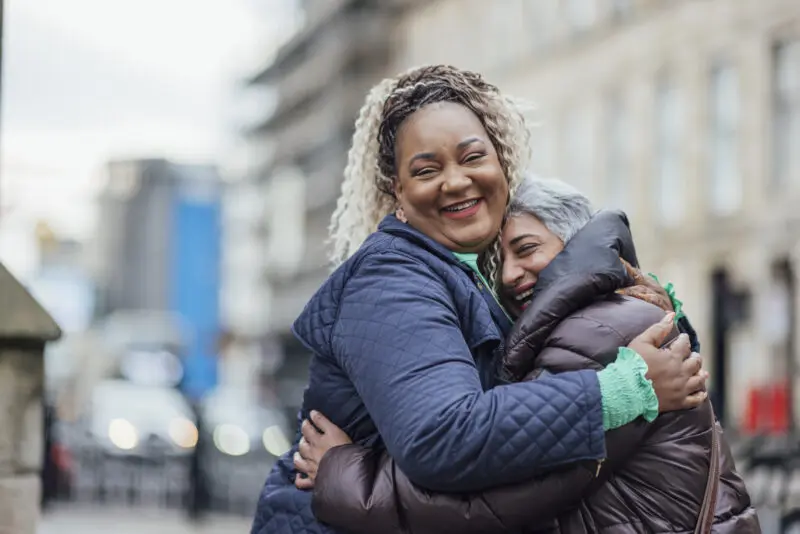 two mid-adult female professionals smiling and embracing in an outdoor urban setting