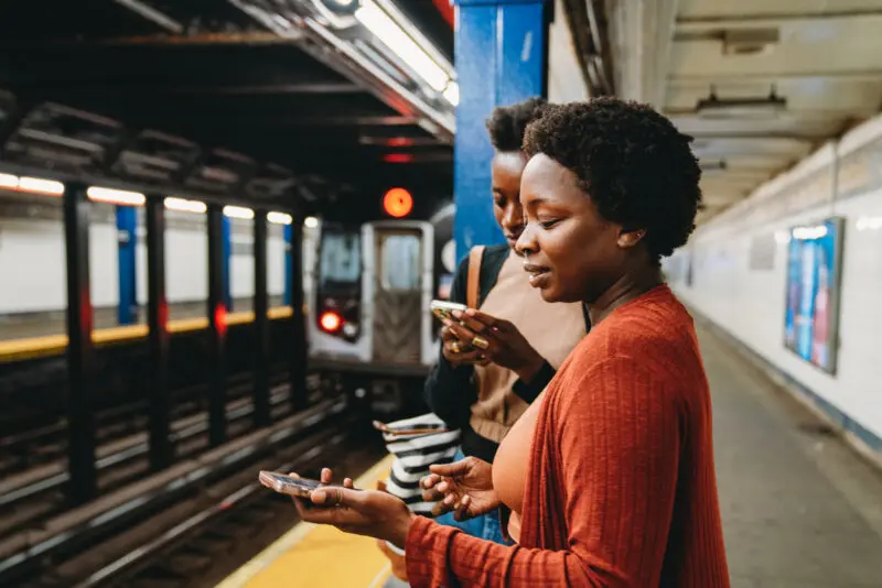 two women looking at their phones while waiting on a subway platform