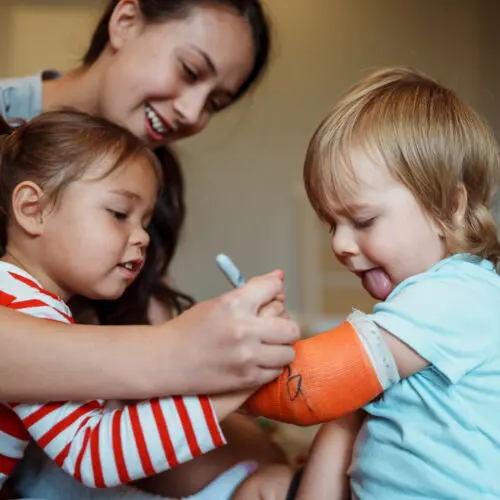 a little boy looks down at his arm in an orange cast while his mother and young sister sign it