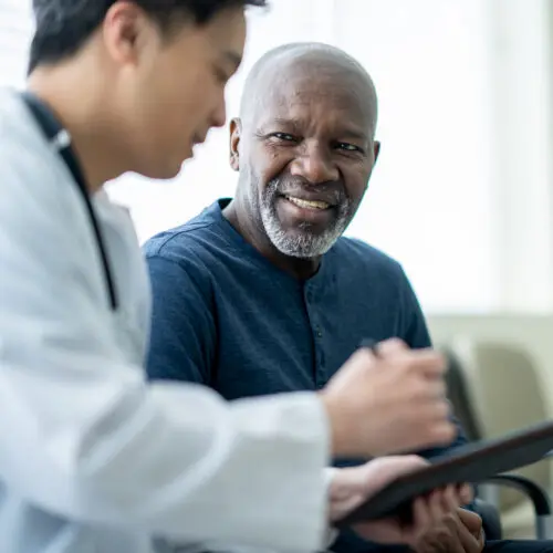 senior gentleman sits in a waiting room beside his doctor as they review test results on a tablet together