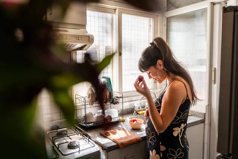 mid adult pregnant woman caressing her belly and eating a strawberry in domestic kitchen