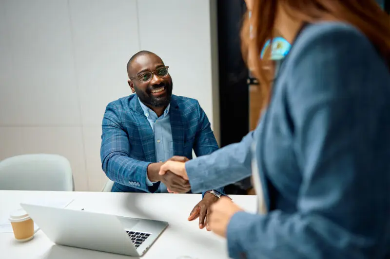man sitting at a white desk with his laptop is looking up and smiling while shaking a woman's hand
